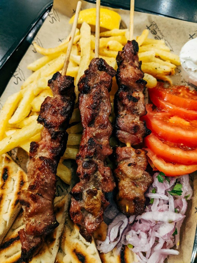 Three grilled meat skewers with french fries, sliced tomatoes, red onions, and pita bread